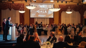 WEHBA Chair's Gala event featuring a speaker at a podium in front of an audience, with a large screen displaying "WEHBA Chair's Gala" and Remington Homes logo, illustrating ROC Media's event production services.