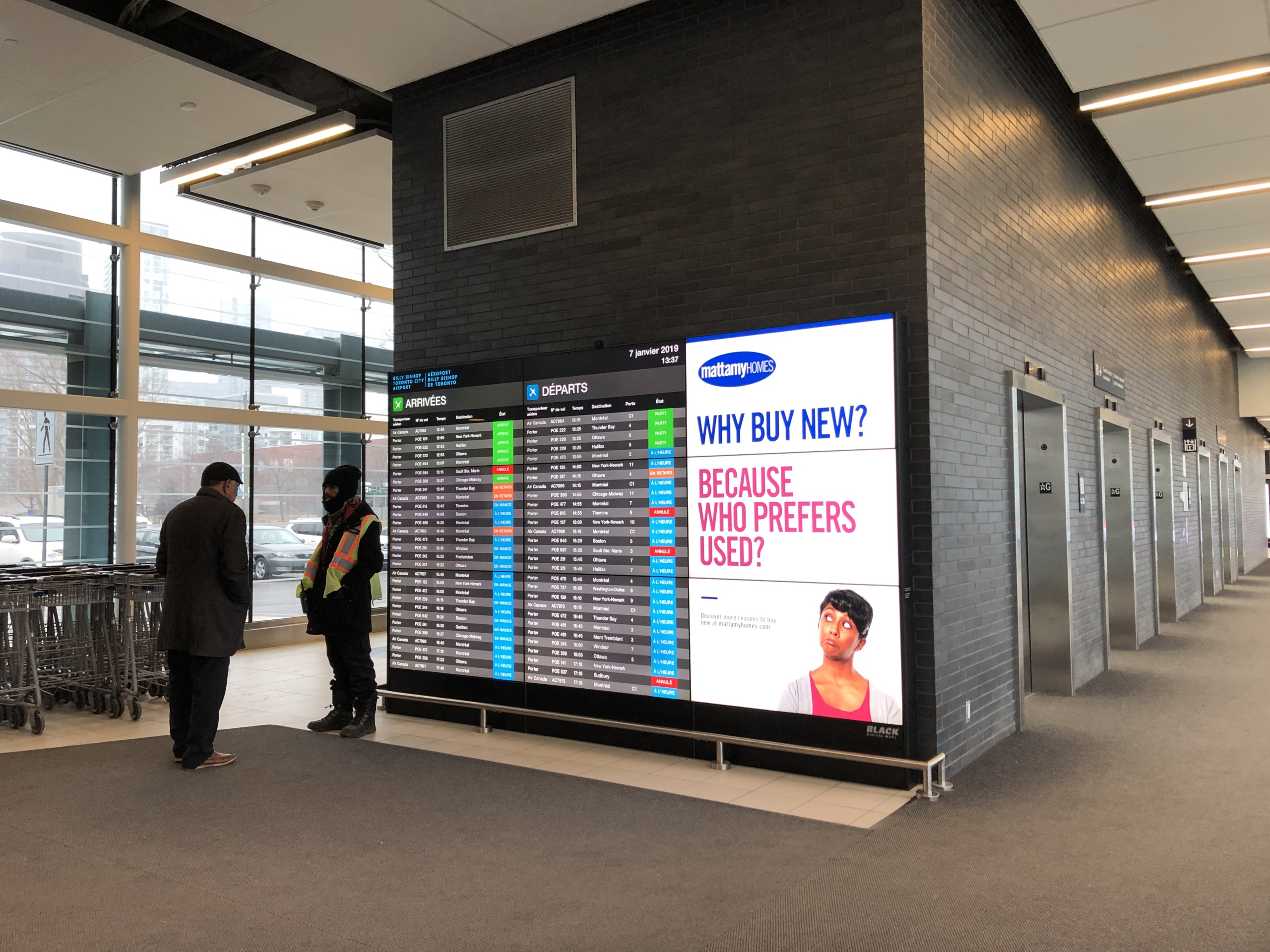 Man in a coat talking to a staff member in a reflective vest near a digital display board showing flight information and a Mattamy Homes advertisement with the text "WHY BUY NEW? BECAUSE WHO PREFERS USED?" in an airport setting.
