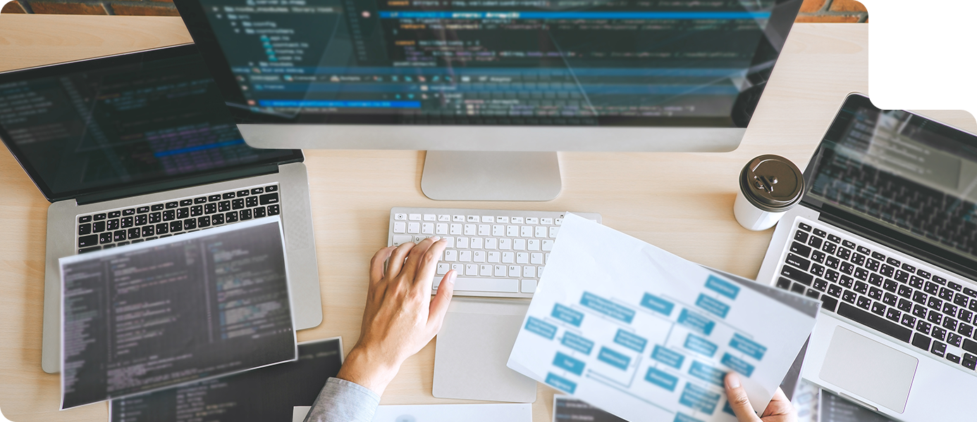 Person typing on a keyboard surrounded by multiple laptops displaying code and a printed document with flowchart elements, illustrating web design and development services offered by ROC Media.