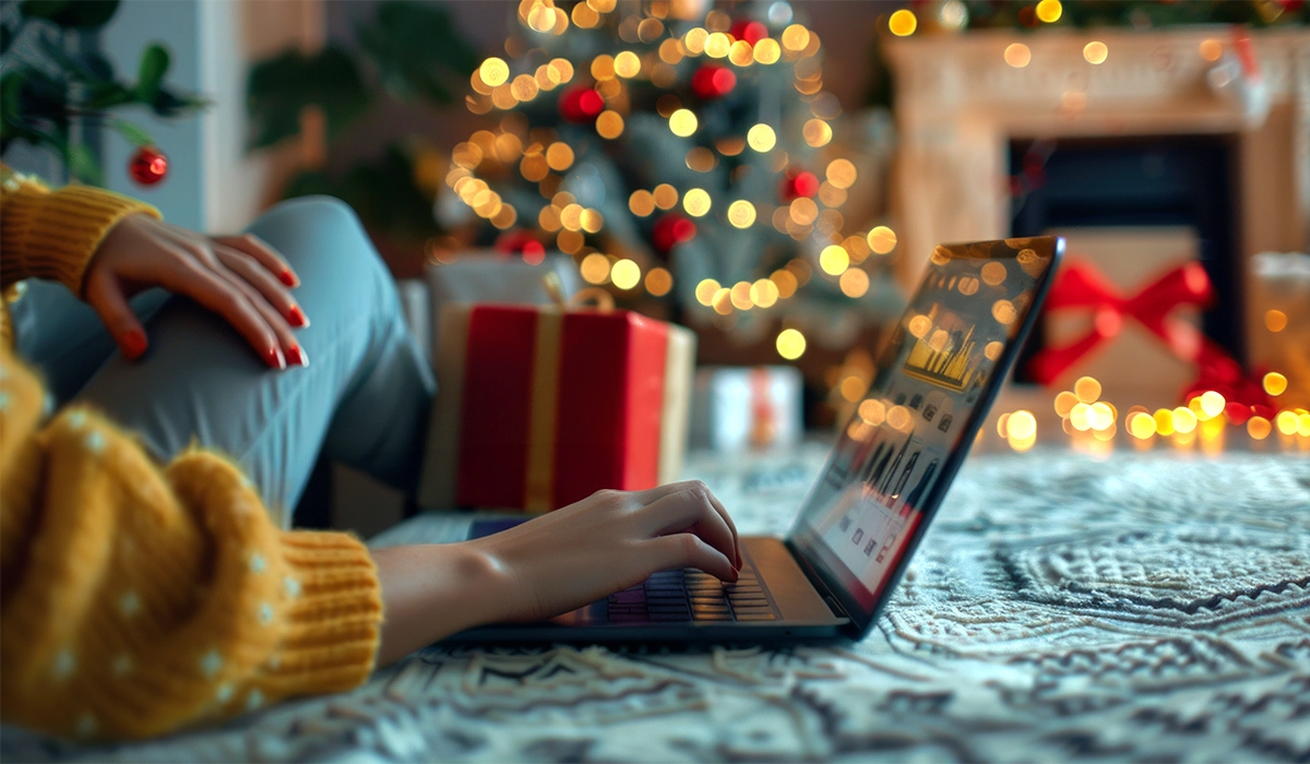 Person using a laptop in a cozy holiday setting with a decorated Christmas tree and wrapped gifts, illustrating the importance of online engagement during the holiday season for lead generation.
