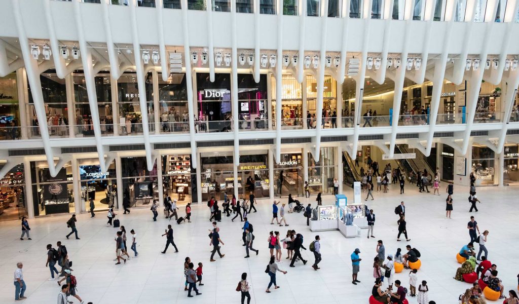 Crowded shopping mall interior with various luxury brand stores, shoppers walking, and modern architectural design elements.