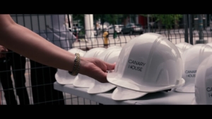Hand reaching for a white hard hat labeled "CANARY HOUSE" among a row of identical helmets, set against a backdrop of a construction site, highlighting the Canary House Penthouse Tour event organized by ROC Media Inc.