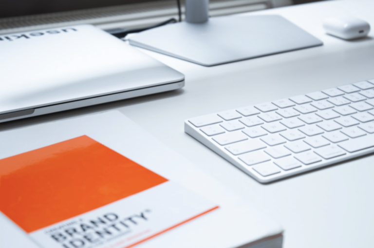 Laptop and keyboard on a white desk with a book titled "BRAND IDENTITY" featuring an orange cover, emphasizing the importance of visual identity in marketing and brand recognition.